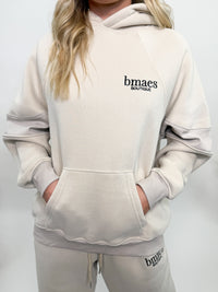 Cream fleece-lined luxury lounge Bmaes hoodie with embroidered Bmaes Boutique logo, drop shoulders, kangaroo pocket, and ribbed details. Perfect for cozy lounging or pairing with the matching joggers for a stylish, warm set from Bmaes Boutique.