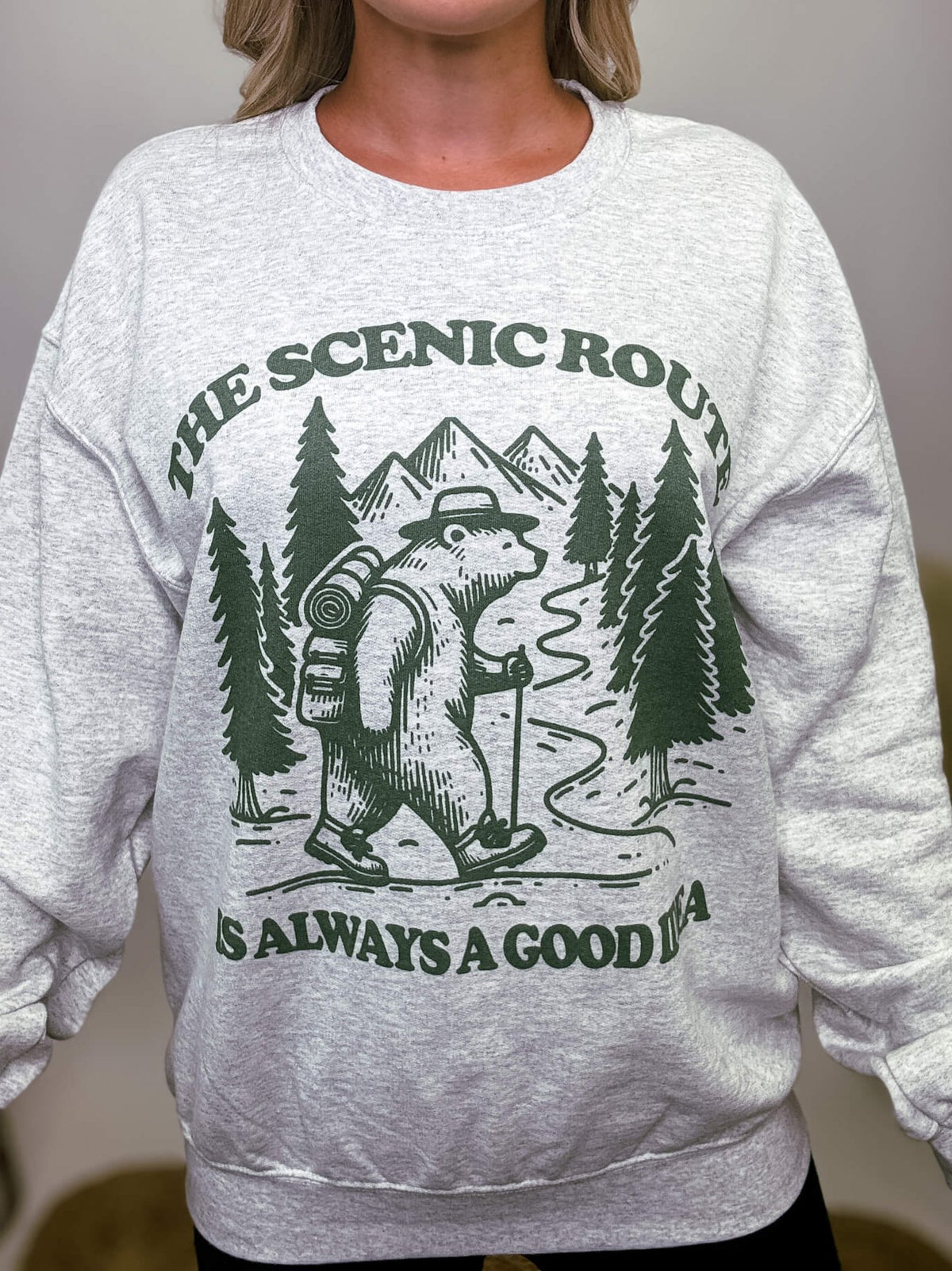 Heather grey fleece lined sweatshirt with a bear hiking graphic and the text 'The scenic route is always a good idea.' Perfect for fall outdoor activities. Model Brooke is 5'4" wearing size small.