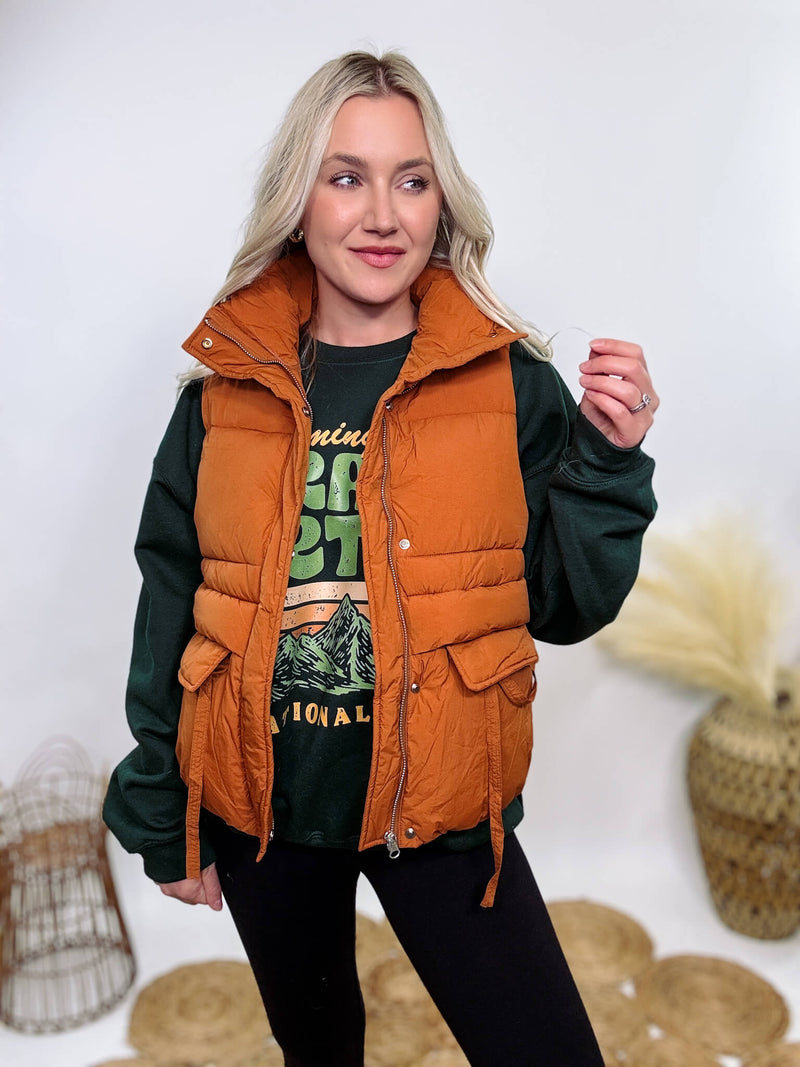 Shop the Rusty Burnt Orange Cargo Puffer Vest at Bmaes Boutique. Made from 100% polyamide, this stylish and versatile vest features two flap pockets and is perfect for layering over sweatshirts or tees.