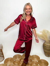 Burgundy satin pajama set featuring a button-down short sleeve shirt and long pants with an elastic waist, true to size relaxed fit. Model Brooke is 5'4" wearing size small.